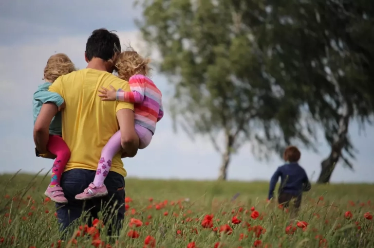 Father carrying two daughters in a field, son running away.