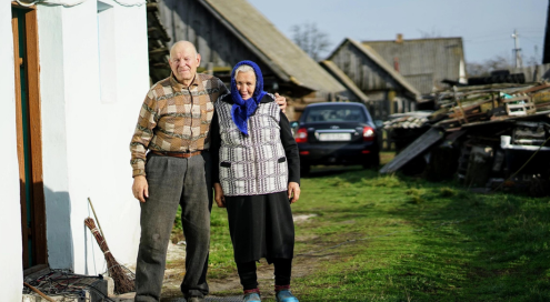 Old couple standing beside a house and near cars.