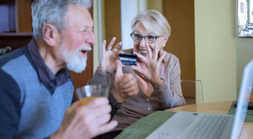 Elderly man happy on computer wife holds credit card.