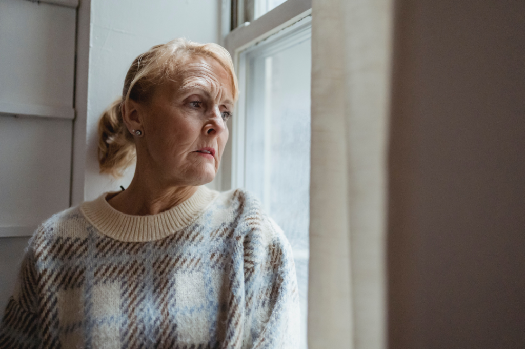 A retired woman staring out of a window with concerns.