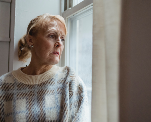 A retired woman staring out of a window with concerns.