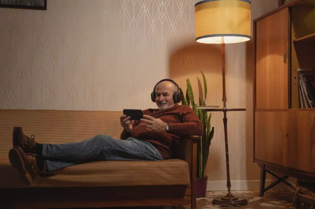 A self-funded retiree sitting on a couch wearing headphones and watching on his smartphone.