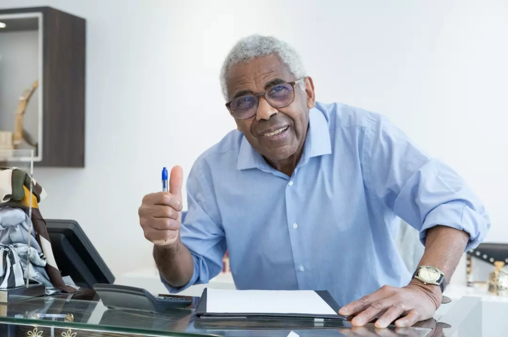 An old man in blue sleeves doing a thumbs up after signing a paper.