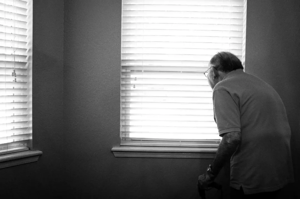 Old man standing and looking from a window inside a small white room.