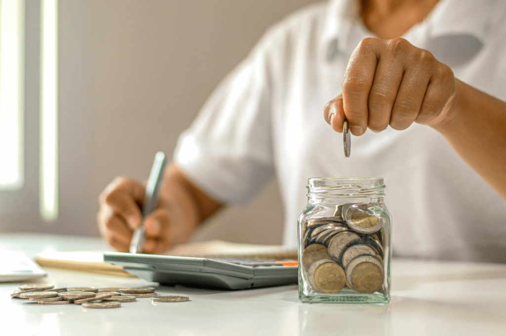 Person in white polo shirt saving coins in a glass of jar filled with coins.
